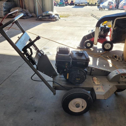 A lawn vacuum machine with a mounted engine on a concrete floor. Nearby, a smaller red and white machine is visible, with bags stacked in the background.