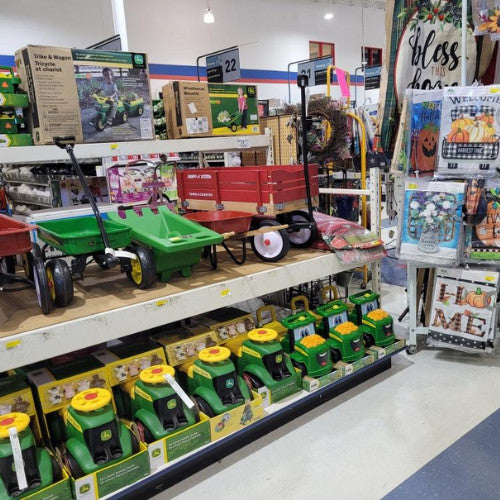 A store aisle displays green toy lawn mowers and red wagons on shelves, with decorative fall-themed signs and wreaths hanging nearby, creating a festive atmosphere.