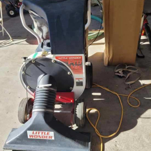 A Little Wonder lawn vacuum with a hose and large nozzle is parked on a concrete floor. A yellow extension cord is coiled beside it, and sunlight casts shadows.