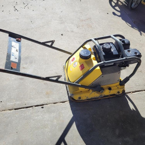 A yellow plate compactor on a concrete surface, casting a shadow. It has a metal handle and visible engine, used for soil compaction in construction.