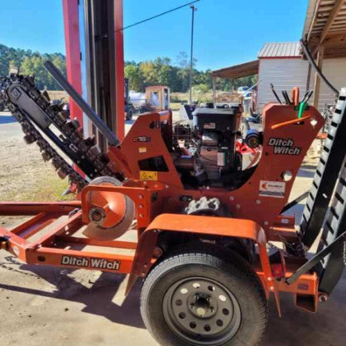A bright orange Ditch Witch trenching machine is parked in a garage, its trencher arm raised. Sunlit trees and a building can be seen outside.